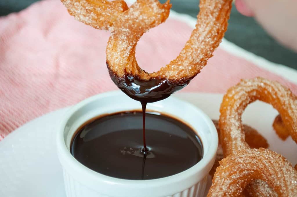 A sugar-coated churro is being dipped into a small white ramekin filled with chocolate sauce, with more churros on the plate in the background.
