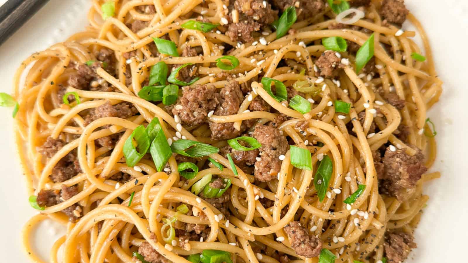 Plate of spaghetti noodles mixed with ground beef, garnished with chopped green onions, black pepper, and sesame seeds.