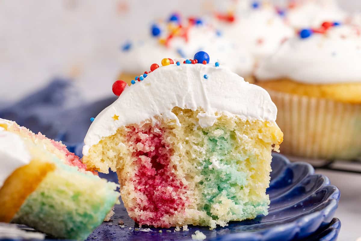 A cupcake with white frosting and colorful sprinkles, cut in half to show rainbow-colored cake inside, sits on a blue plate.