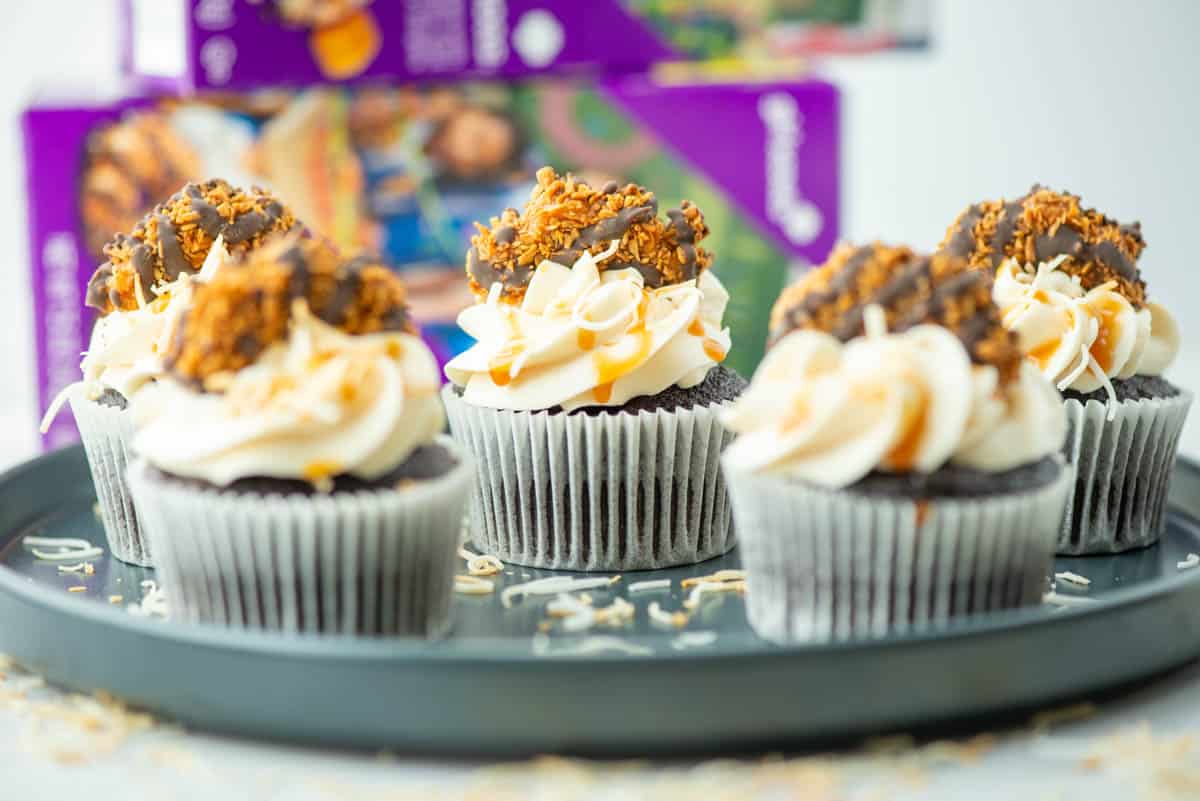 Four chocolate cupcakes with white frosting, topped with cookie pieces and caramel drizzle, are arranged on a round tray. Cookie boxes are visible in the background.