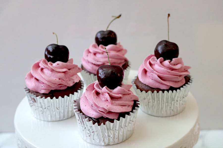 Four chocolate cupcakes in silver wrappers, topped with pink frosting and a whole cherry, displayed on a white surface.
