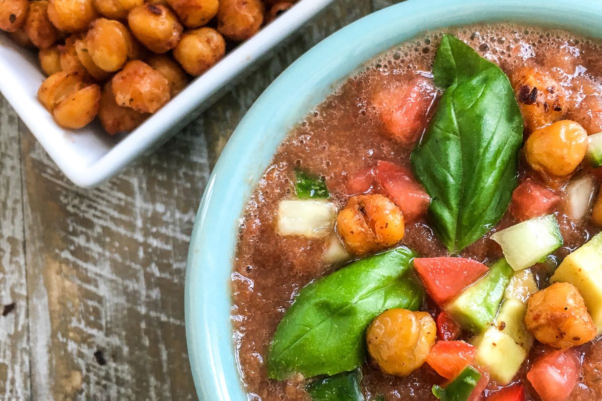 Close-up of a bowl of gazpacho topped with avocado, tomato, cucumbers, basil leaves, and roasted chickpeas, with a dish of extra chickpeas in the background on a wooden surface.