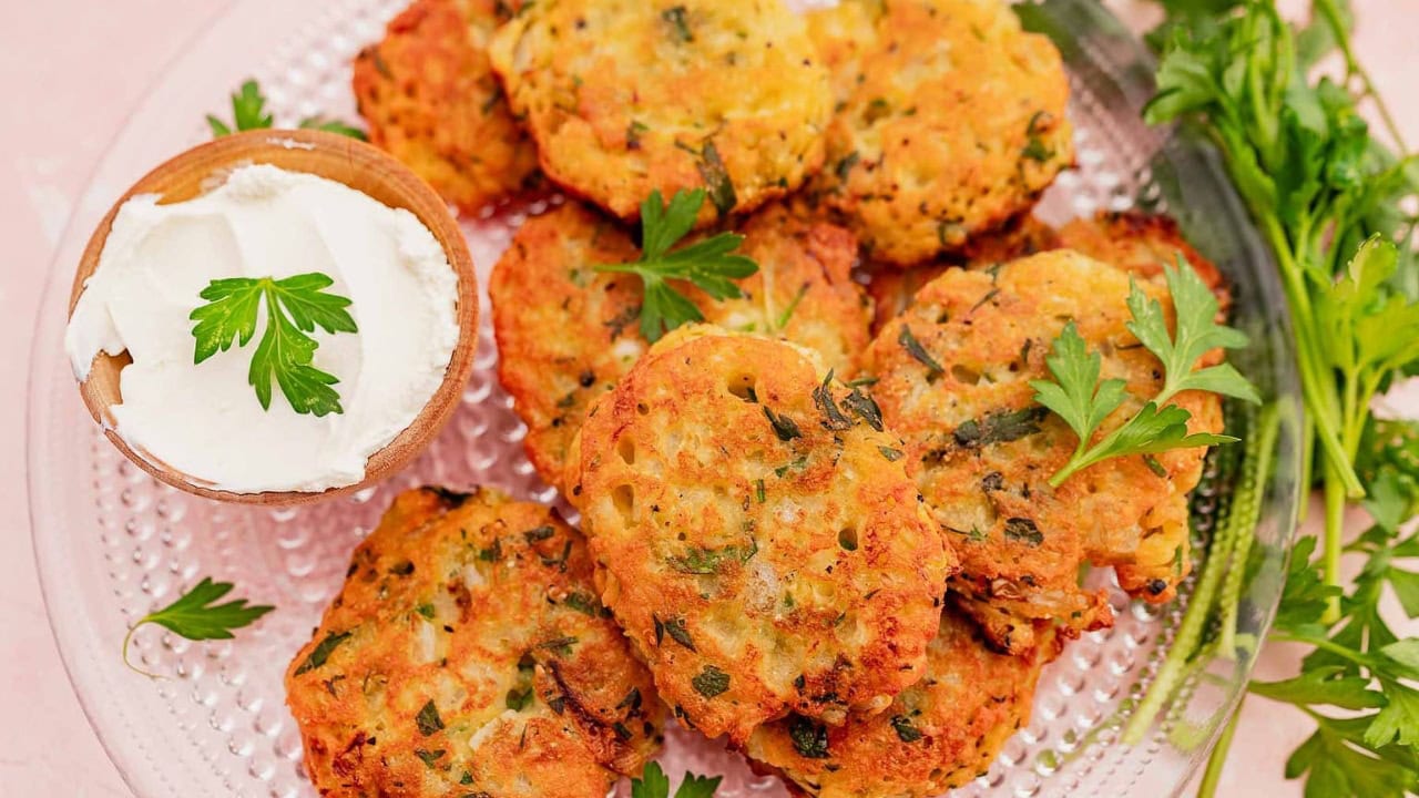 A plate of golden-brown fritters garnished with parsley, served with a small bowl of sour cream and fresh herbs on the side.