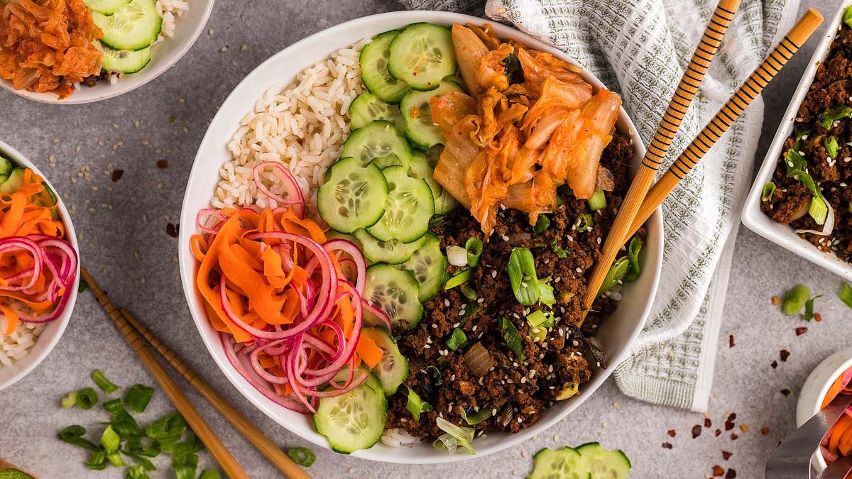 A bowl with rice, sliced cucumbers, pickled carrots and onions, cooked ground meat, kimchi, and chopped green onions, with chopsticks resting on the side.
