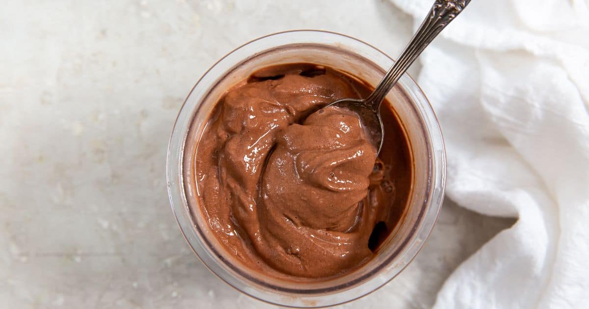 A glass bowl filled with creamy chocolate mousse, with a spoon inside, placed on a light-colored surface next to a white cloth.