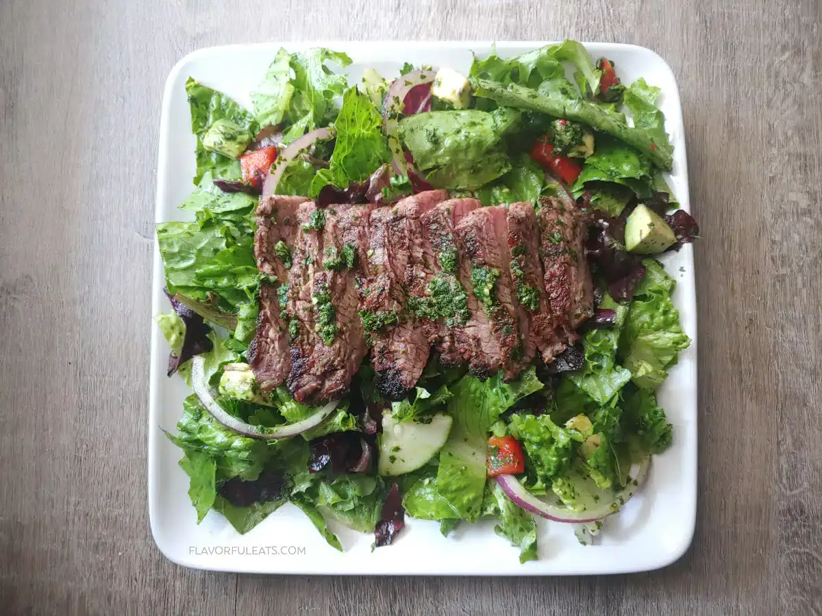 A plate of mixed green salad topped with sliced grilled steak and chopped herbs, served on a square white dish on a wooden surface.