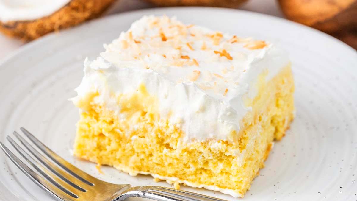 A slice of yellow cake topped with white whipped cream and toasted coconut flakes on a white plate with a fork.