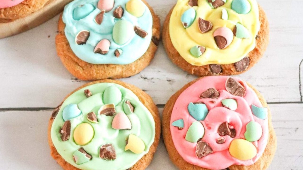 Four cookies with pastel-colored icing in blue, yellow, green, and pink, each topped with pieces of candy-coated chocolate eggs.
