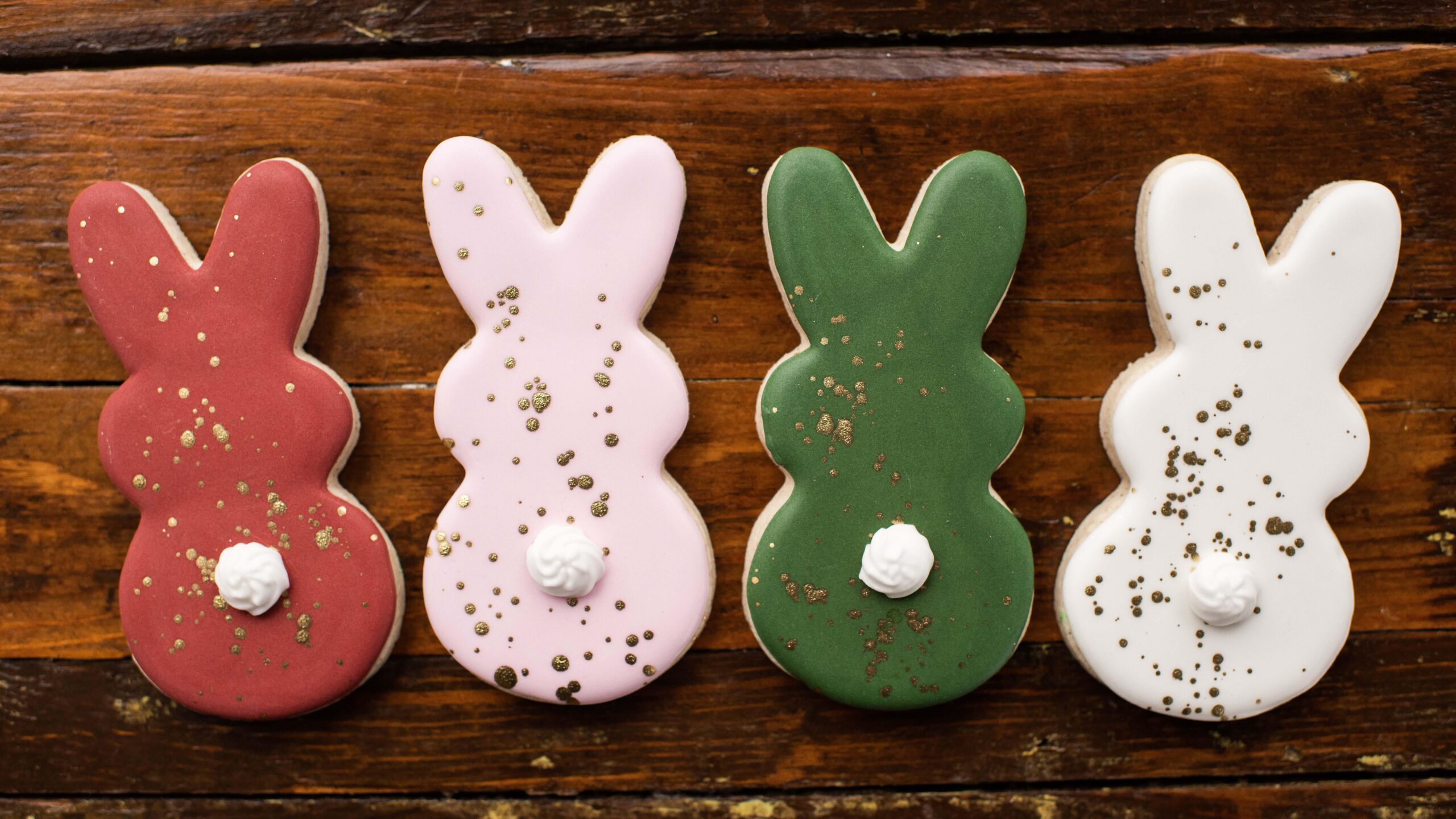 Four bunny-shaped cookies in red, pink, green, and white with gold speckles and white tails, arranged in a row on a wooden surface.