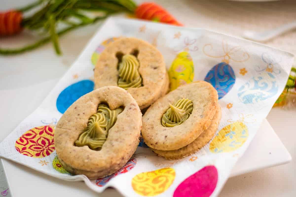 Three oval sandwich cookies with bunny-shaped cutouts and green filling are placed on a napkin decorated with colorful egg patterns.