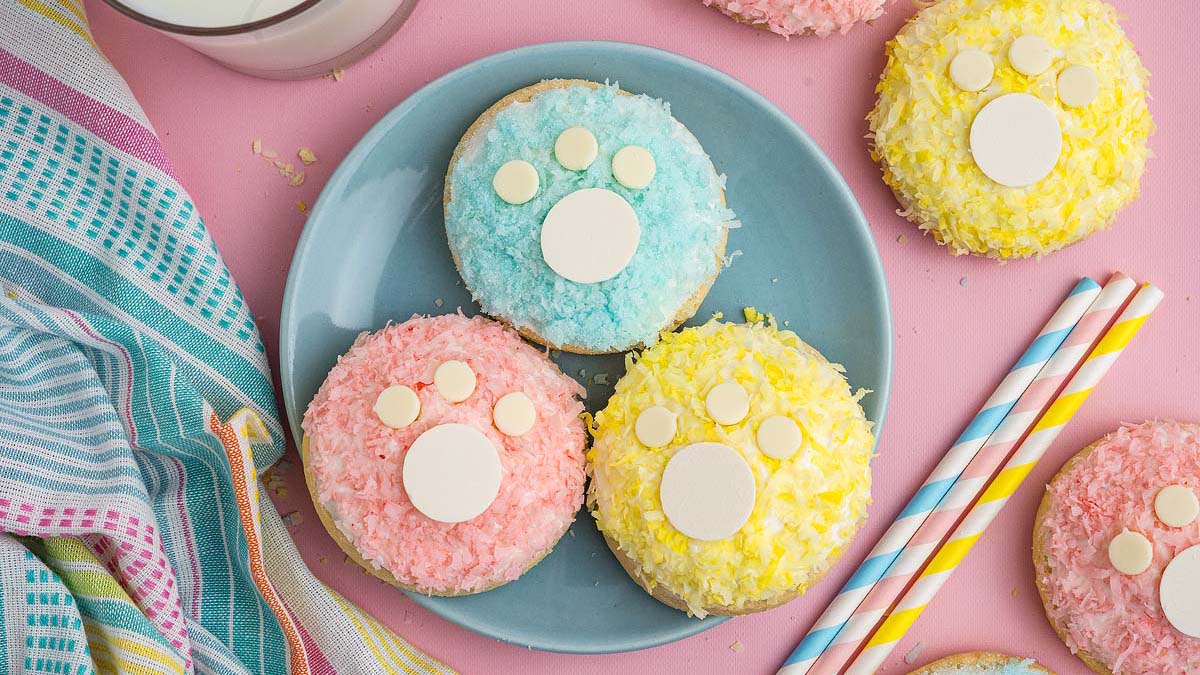 Three cookies with pastel-colored coconut shreds and white chocolate chips are arranged on a blue plate, resembling animal paw prints; striped straws and a towel are nearby.
