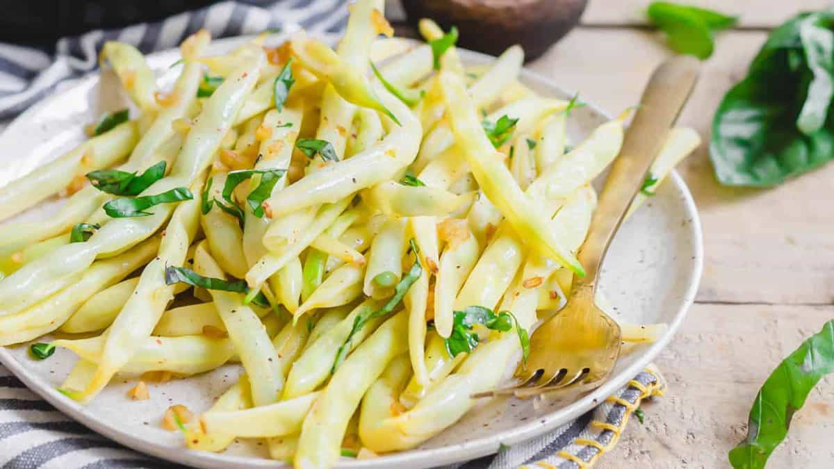 A plate of cooked yellow wax beans garnished with chopped herbs, served with a gold fork on a striped napkin.