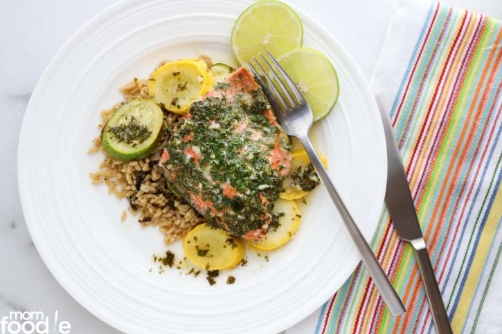 A plate with baked herb-crusted salmon, sliced yellow squash and zucchini, brown rice, and lime slices, with a fork and knife on the side next to a striped napkin.
