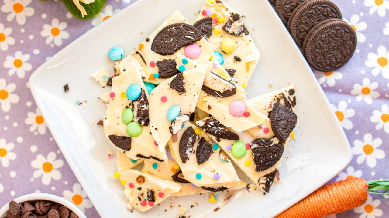 White chocolate bark topped with crushed chocolate sandwich cookies and colorful candy pieces on a white plate, placed on a floral-patterned cloth.