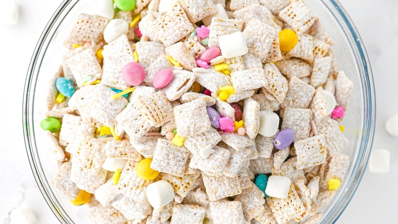 A glass bowl filled with powdered cereal squares, mini marshmallows, and colorful candy pieces on a white surface.