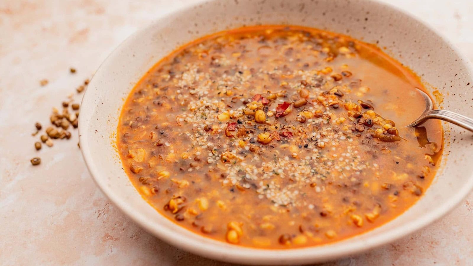 A bowl of lentil soup with visible lentils, seeds, and a light broth, served with a spoon on the side of the bowl.