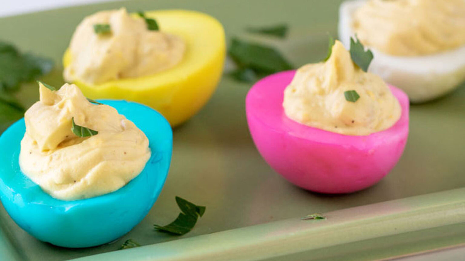 Three deviled eggs with dyed egg whites in blue, yellow, and pink, filled with creamy yolk mixture and garnished with chopped herbs on a green tray.