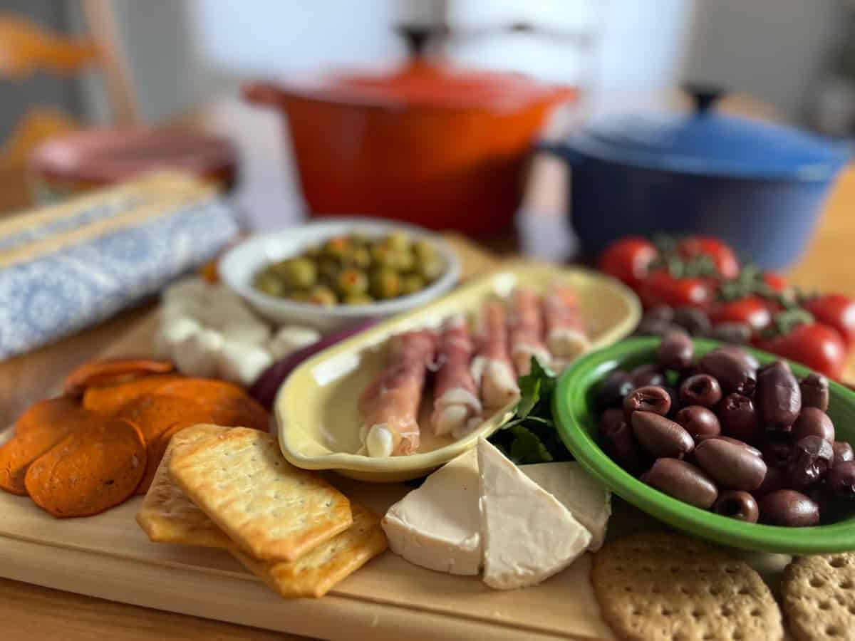A wooden board with assorted crackers, cheeses, cured meats, olives, tomatoes, and two covered pots in the background.