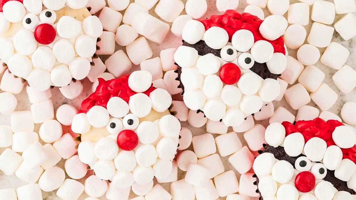 Four cupcakes decorated as Santa faces with red icing and mini marshmallows, surrounded by loose mini marshmallows.