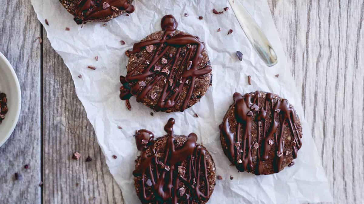 Four round chocolate cookies with chocolate drizzle and cocoa nibs rest on crumpled parchment paper atop a wooden surface, with a knife nearby.