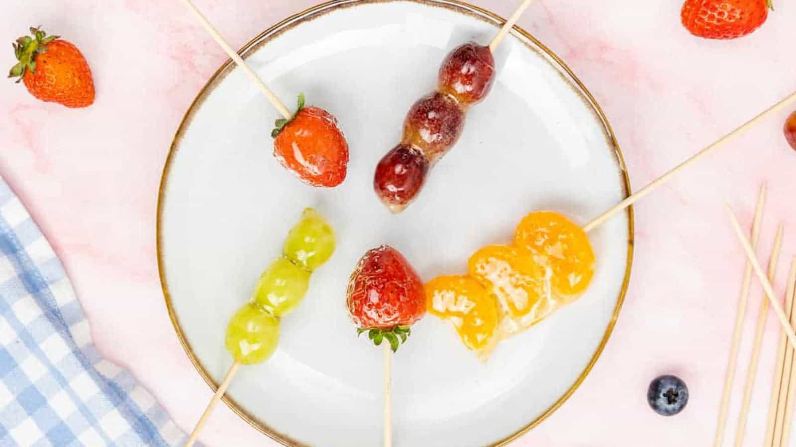Five fruit skewers with strawberries, grapes, and orange segments on a white plate, set on a pink surface with extra fruit and wooden sticks nearby.