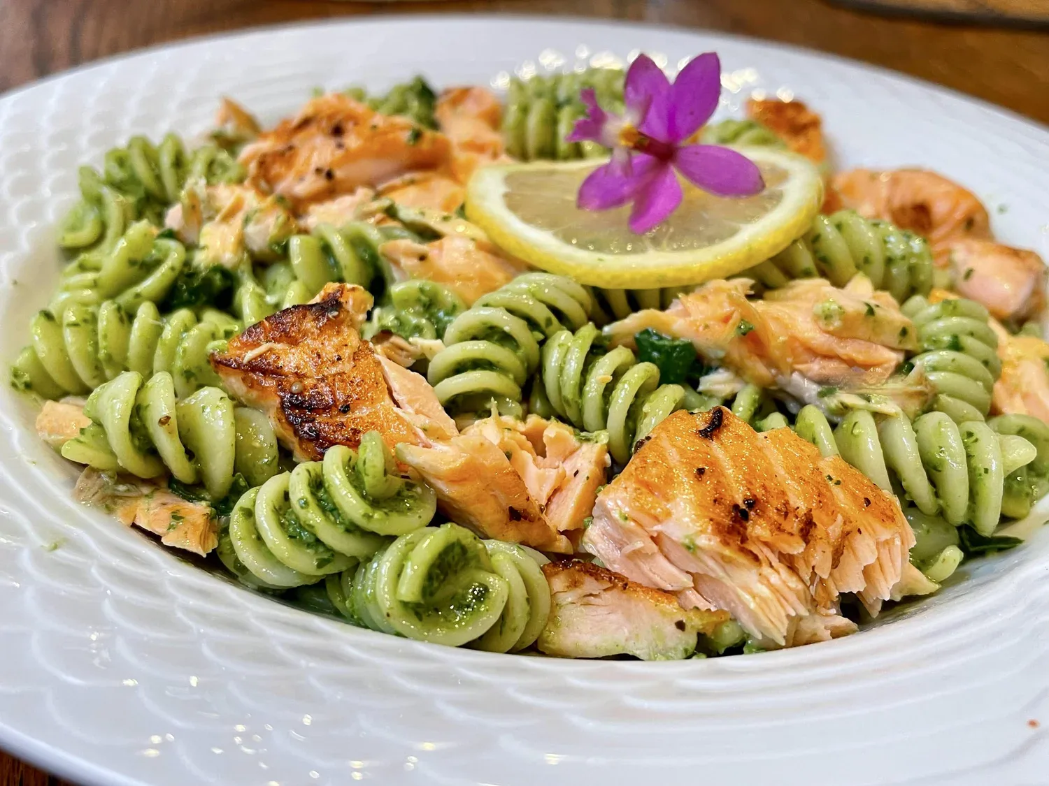 A plate of pesto rotini pasta topped with grilled salmon pieces, a lemon slice, and a small purple flower garnish.