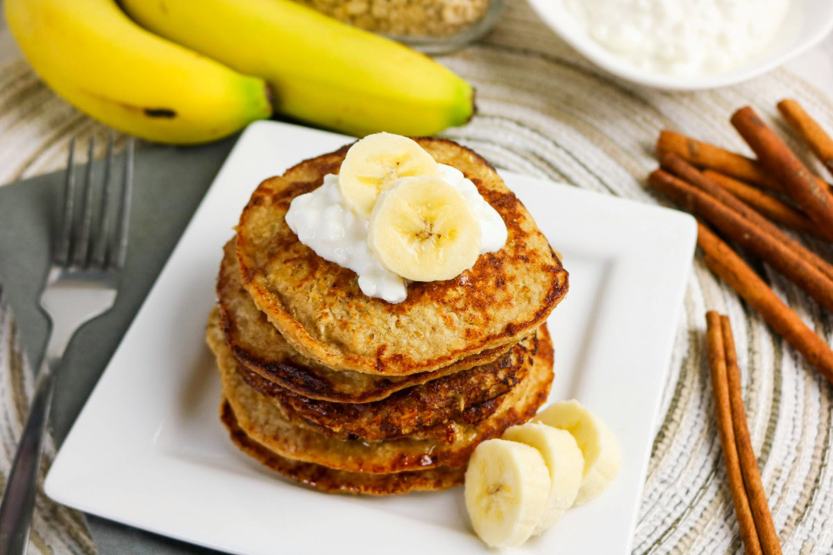A stack of pancakes topped with banana slices and a dollop of cottage cheese on a white plate, with bananas and cinnamon sticks in the background.