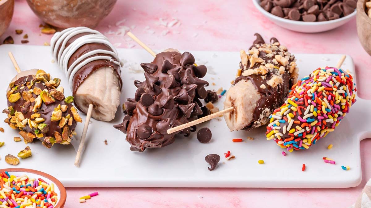 Four chocolate-covered banana pops on sticks, each decorated with toppings: chopped pistachios, white drizzle, chocolate chips, nuts, and rainbow sprinkles, placed on a white tray.