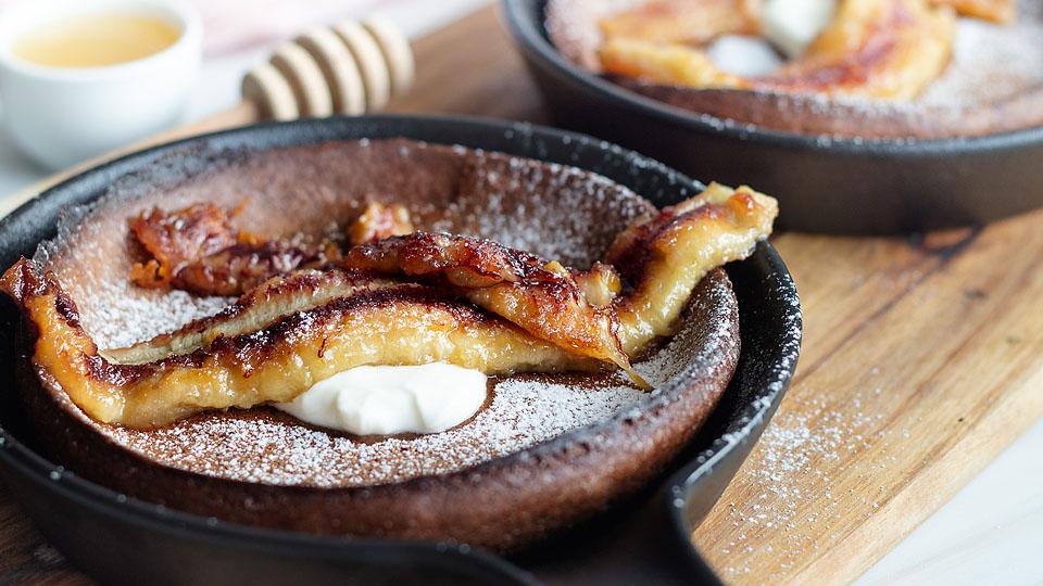 A cast iron skillet with a baked pancake topped with caramelized banana, powdered sugar, and a dollop of cream, set on a wooden board.