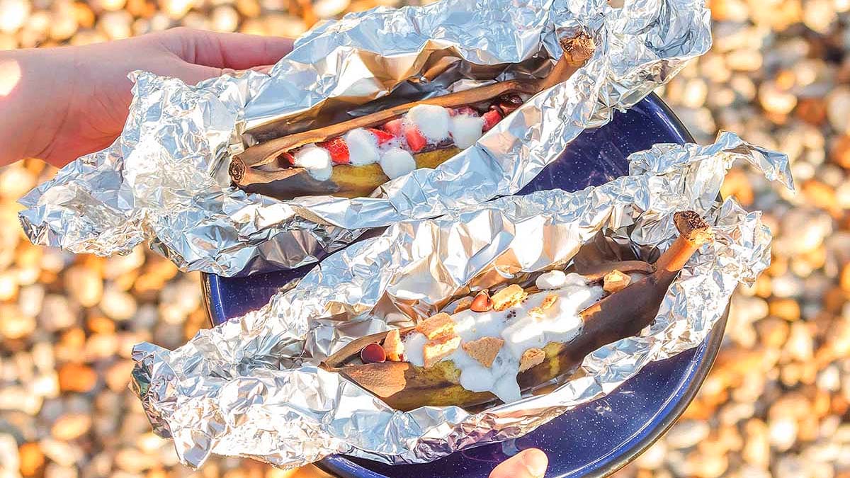 Two bananas in foil, each split and filled with marshmallows, chocolate chips, and strawberries, placed on a blue plate held outdoors.