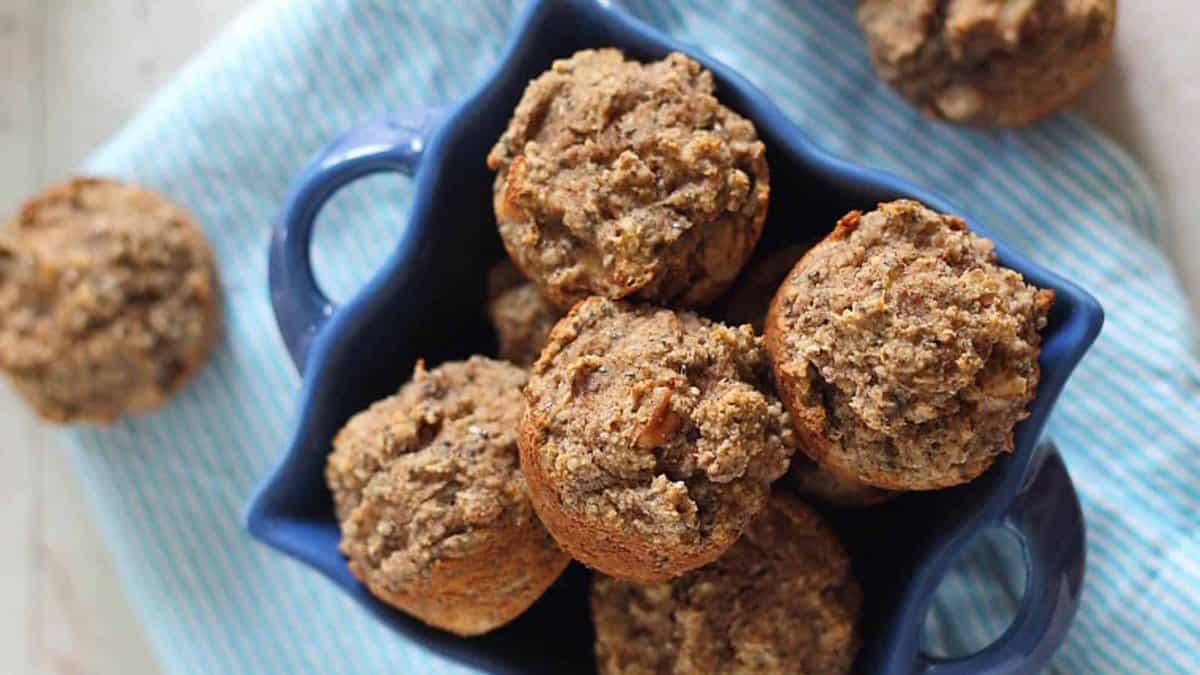 A blue dish filled with several brown, crumbly muffins sits on a blue and white striped cloth; two additional muffins are nearby.