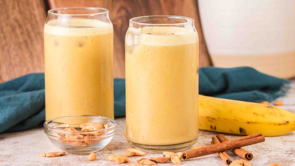 Two glasses of yellow smoothie are placed on a table with a banana, walnuts, and cinnamon sticks beside them.