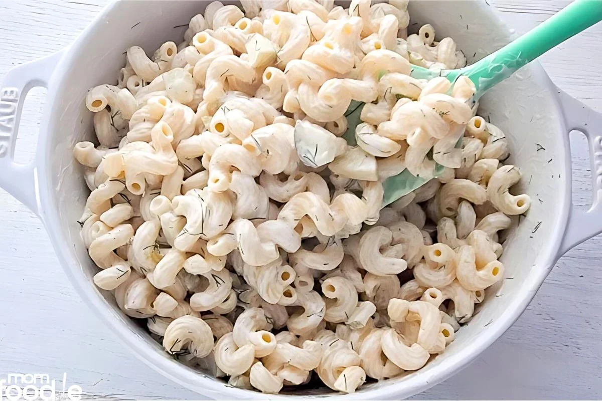A white bowl filled with cooked cavatappi pasta coated in a creamy white sauce, being stirred with a green spatula.