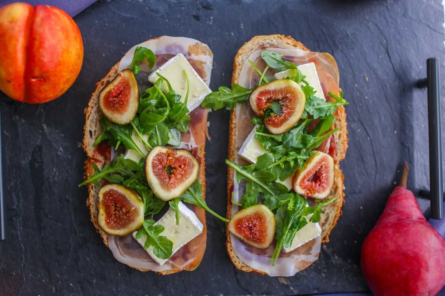 Two slices of bread topped with prosciutto, brie cheese, arugula, and sliced figs on a slate board, with a peach and a pear nearby.