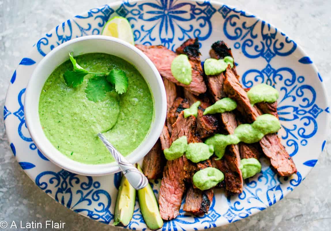 Grilled steak slices topped with green sauce, served on a blue and white patterned plate with a bowl of green sauce, avocado slices, and a lime wedge.