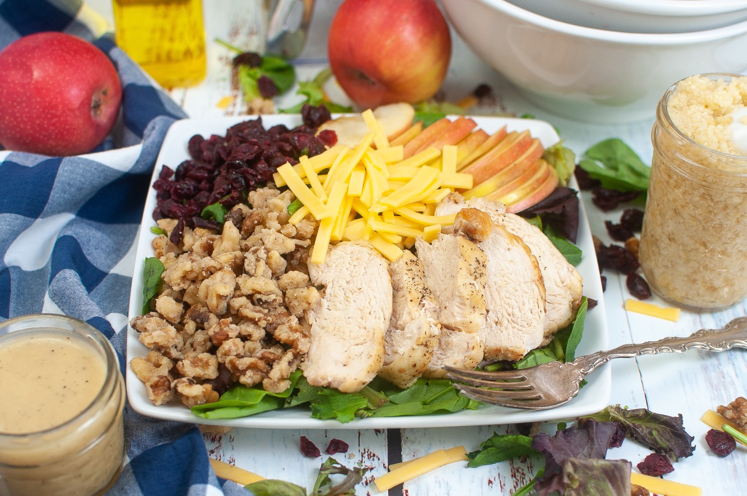 A plate with sliced grilled chicken, shredded cheese, apple slices, walnuts, dried cranberries, and mixed greens, surrounded by dressing, apples, and a jar of shredded cheese.