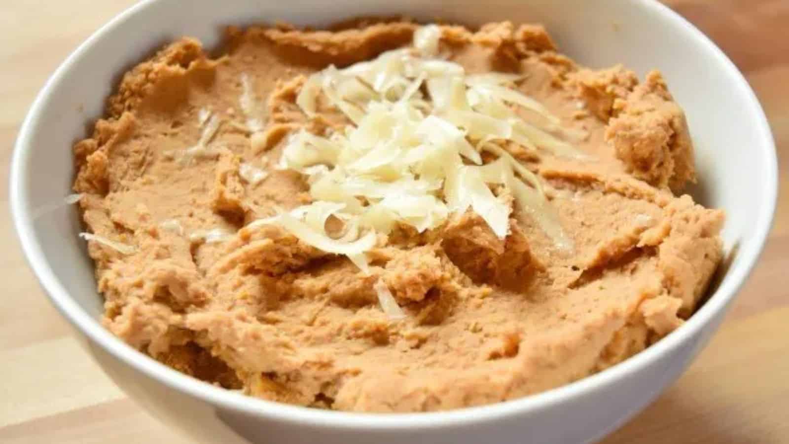 A white bowl filled with refried beans, topped with shredded cheese, sits on a wooden surface.