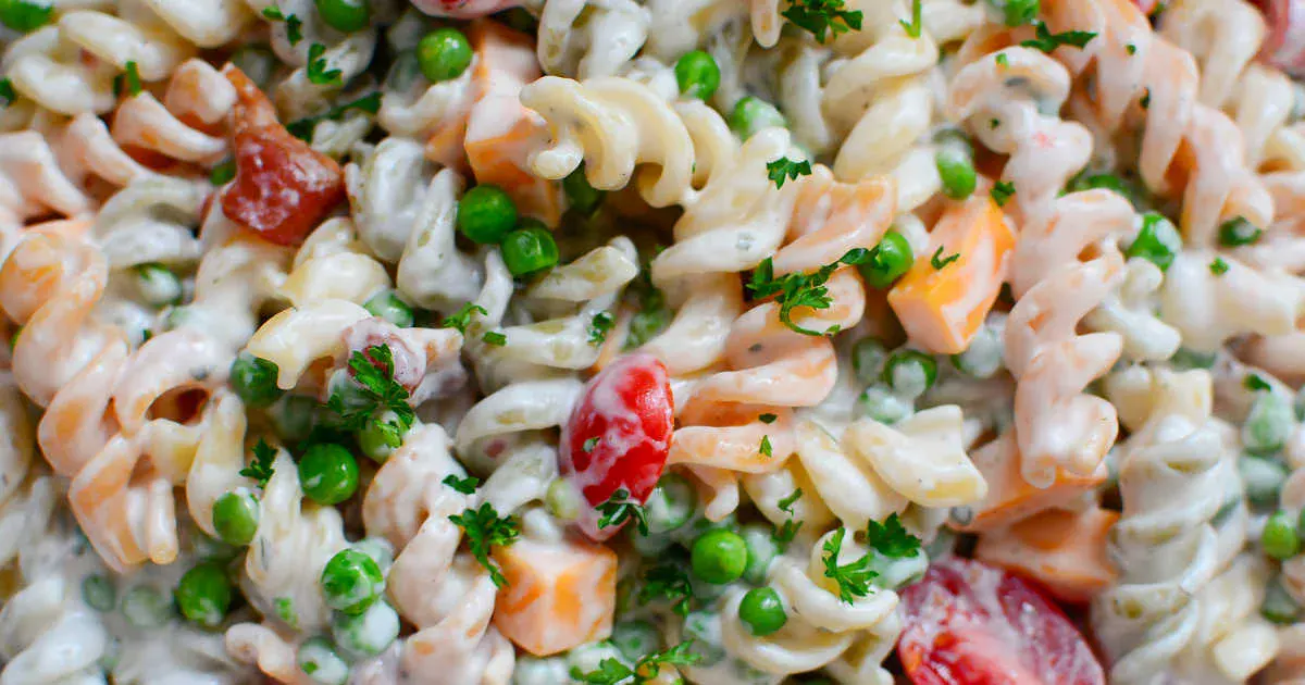 Close-up of creamy pasta salad with rotini noodles, green peas, diced cheese, cherry tomatoes, and chopped parsley.