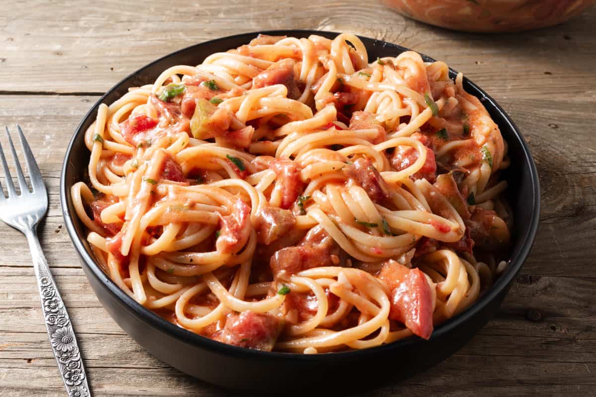 A black bowl filled with spaghetti topped with creamy tomato sauce and herbs sits on a wooden table next to a fork.