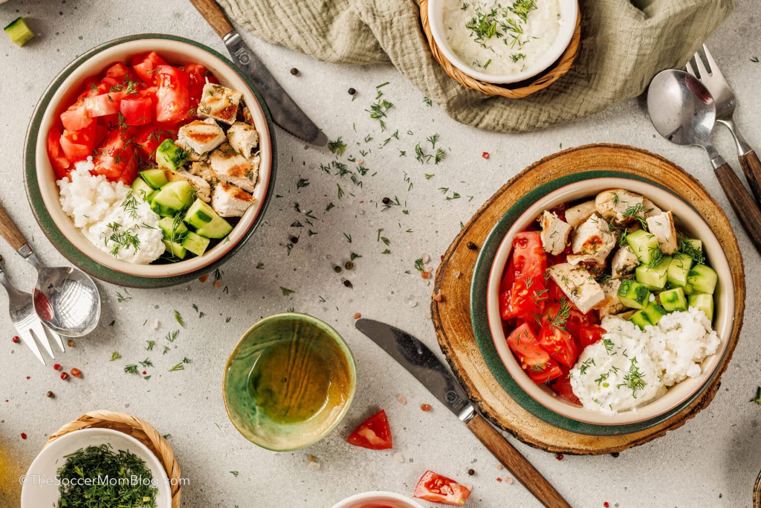 Two bowls filled with chopped tomatoes, cucumbers, grilled chicken, white cheese, and herbs sit on a table with utensils, a cup, and small dishes nearby.