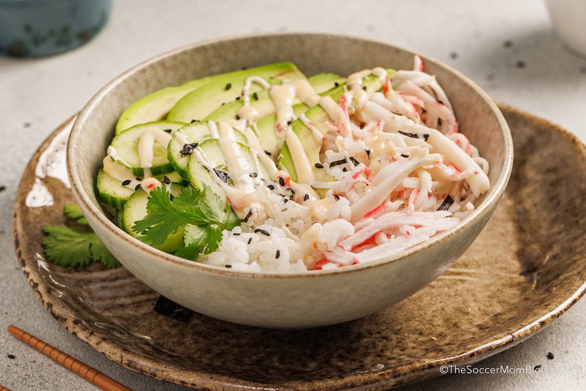A bowl with rice, shredded imitation crab, sliced cucumber, cilantro, black sesame seeds, and a drizzle of creamy sauce.