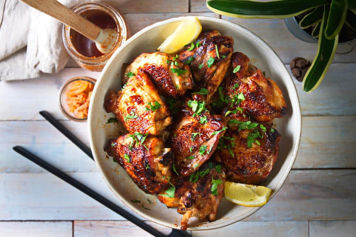 A bowl of grilled chicken garnished with herbs and lemon wedges, with chopsticks, sauce, and a plant visible on a light wooden table.