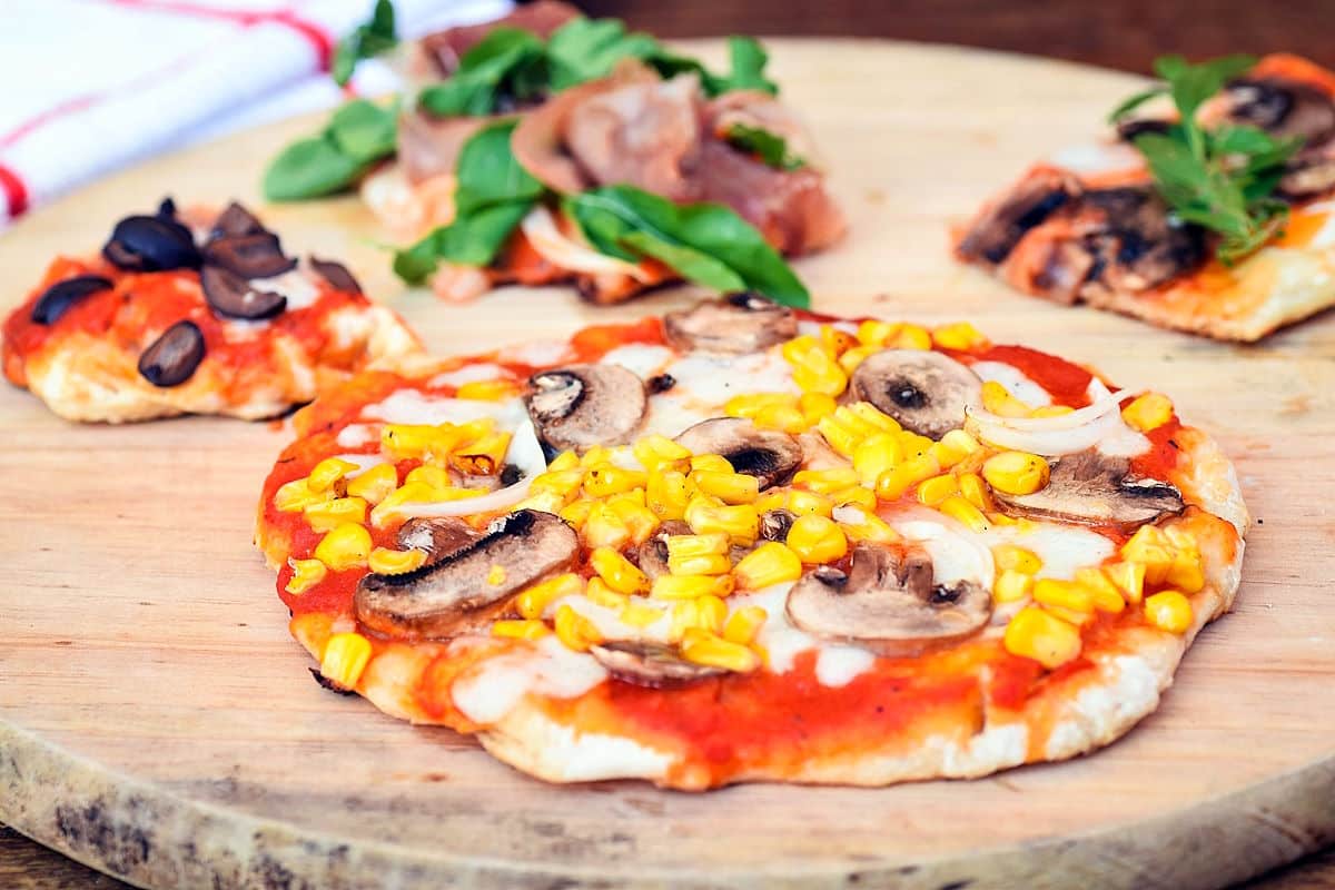 Personal-sized pizza topped with corn, mushrooms, and onions on a wooden board, with two slices of another pizza and greens in the background.