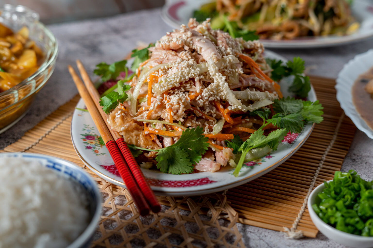 A plate of shredded chicken salad with carrots, cilantro, and sesame seeds, served with red chopsticks and sides of rice and chopped green onions.