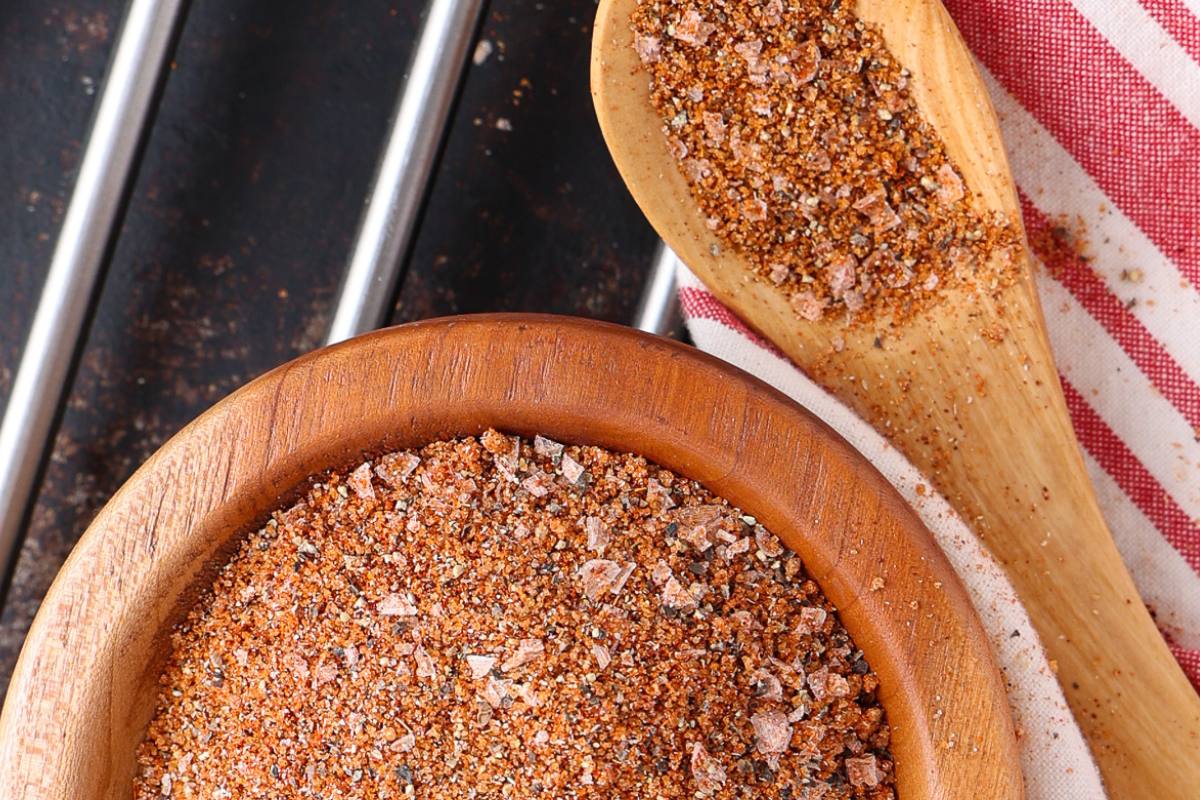 A wooden bowl and spoon filled with a coarse, reddish-brown spice blend sit on a striped cloth near metal grill bars.
