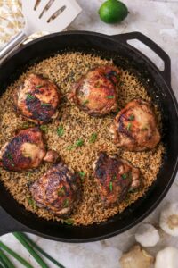A cast iron skillet filled with Jerk Chicken and Rice features six browned chicken thighs, garnished with chopped green onions. Kitchen utensils, lime, garlic, and chives surround the skillet.