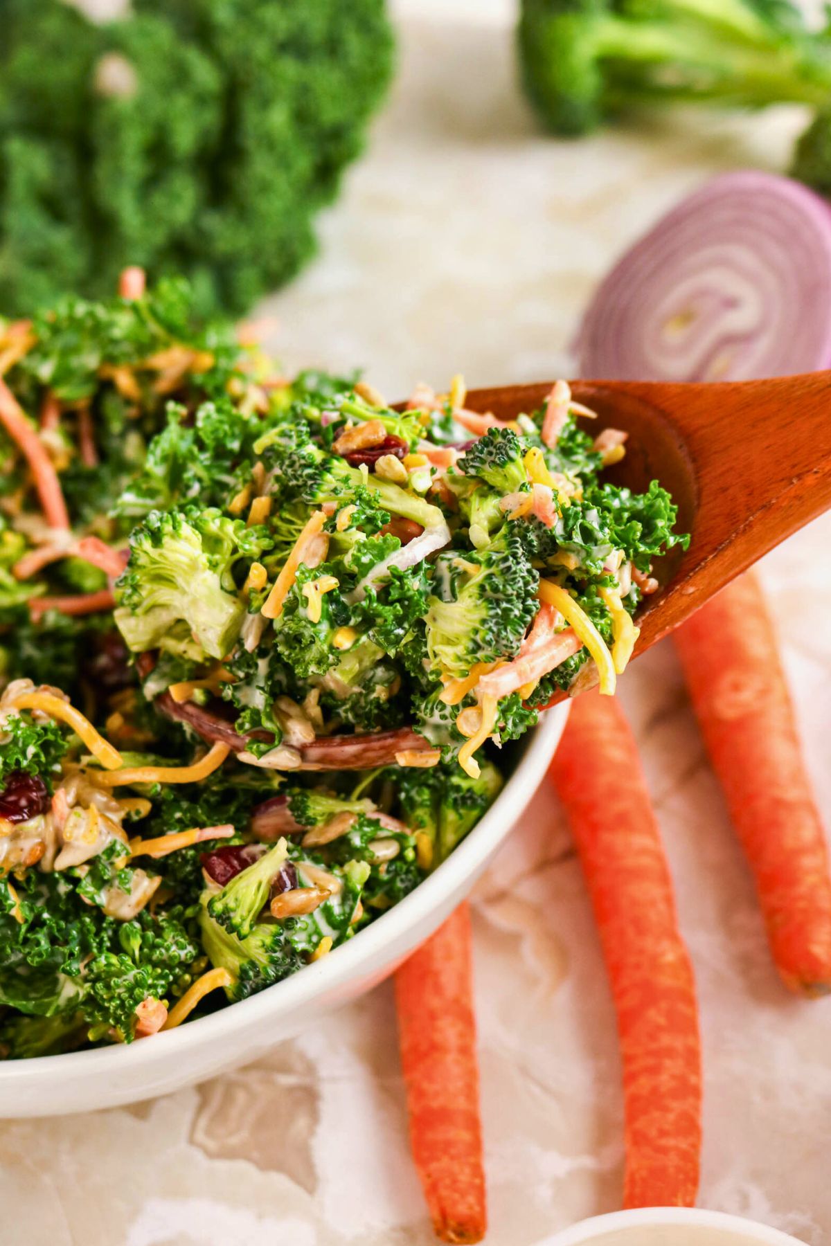 A wooden spoon lifts a portion of broccoli salad with shredded carrots, cheese, and greens from a white bowl. Whole carrots, onion, and broccoli are visible in the background.