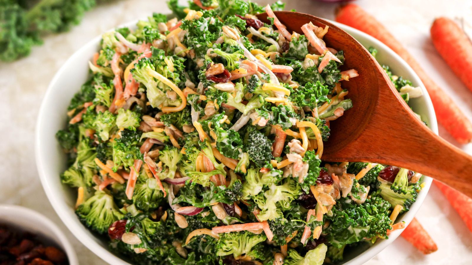 A bowl of broccoli salad with shredded carrots, cheese, cranberries, and seeds, being stirred with a wooden spoon.