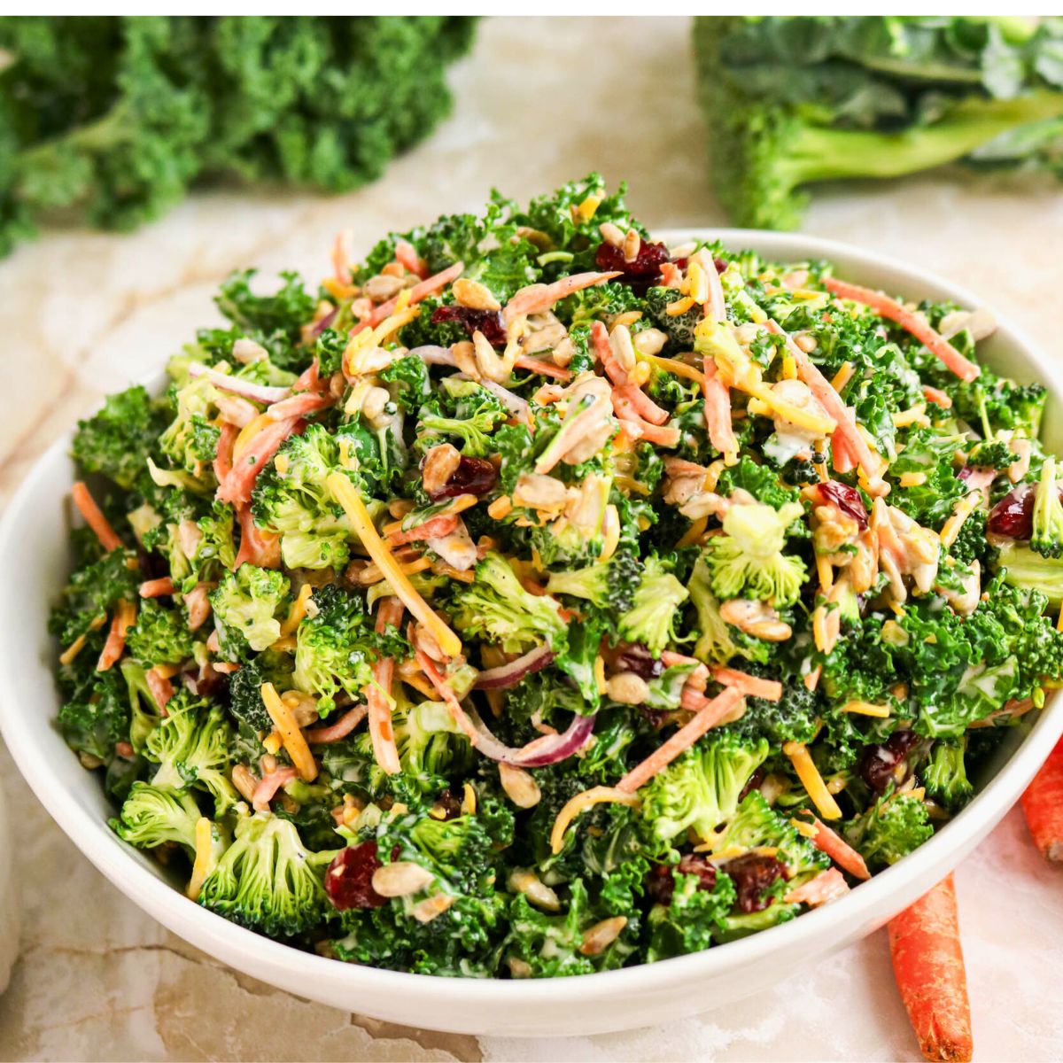 A white bowl filled with a broccoli salad containing shredded carrots, red onion, sunflower seeds, and cranberries, set on a marble surface with fresh greens in the background.