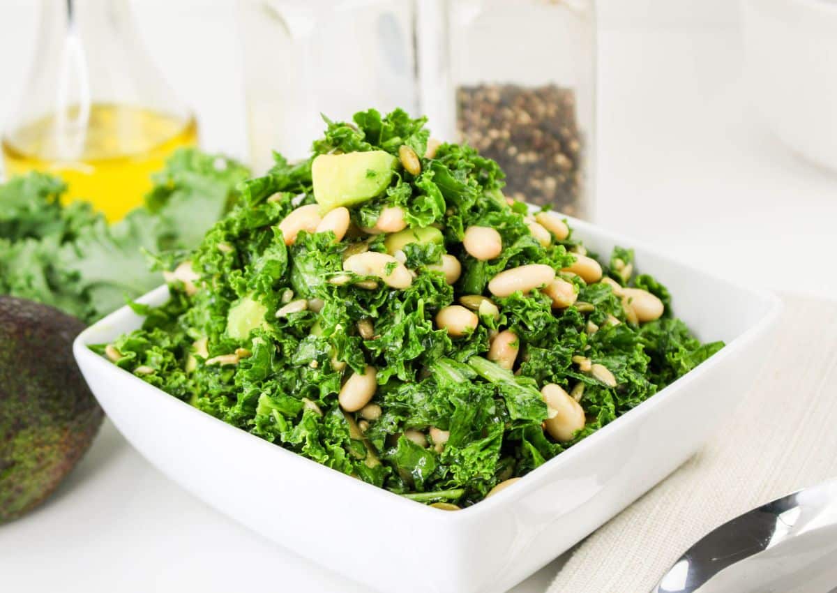 A square white bowl filled with a kale salad topped with white beans, avocado pieces, and seeds, with bottles and kale leaves in the background.